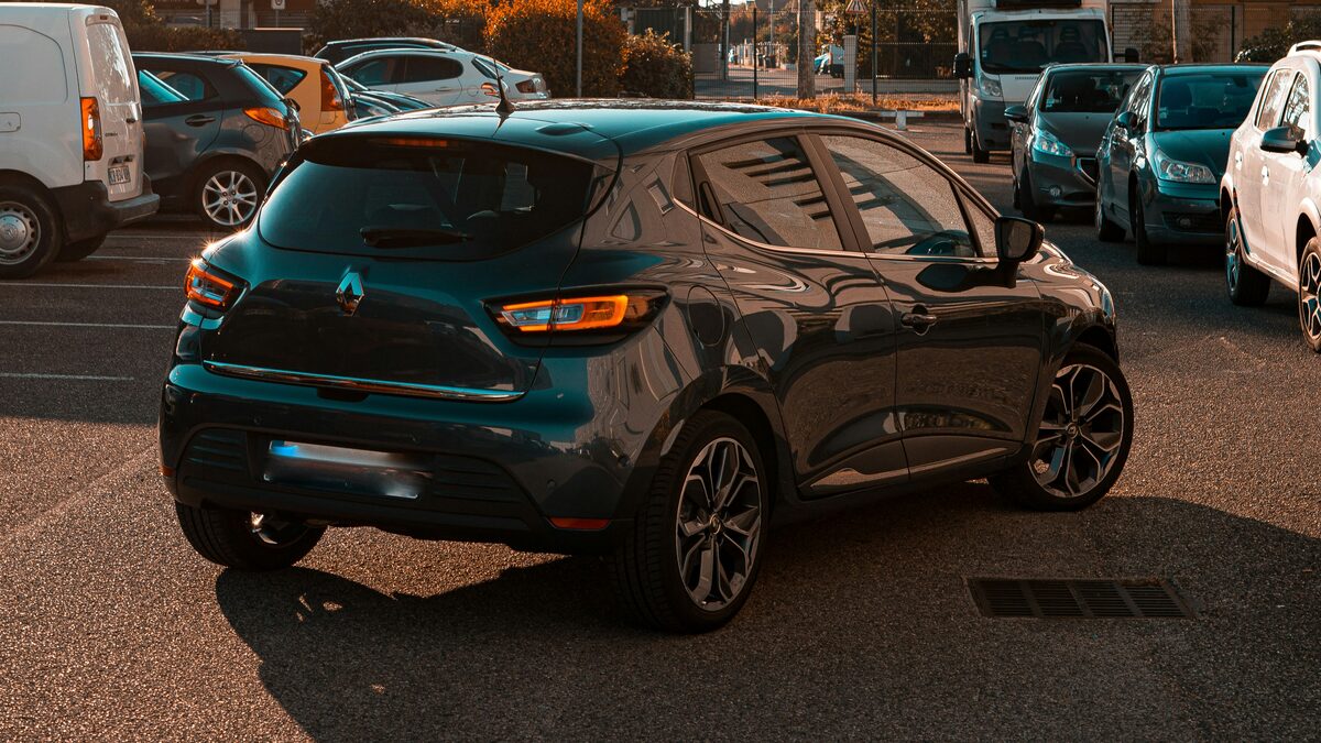 A teal hatchback photographed in a busy car park at sunset, hemmed in by other parked cars and a van